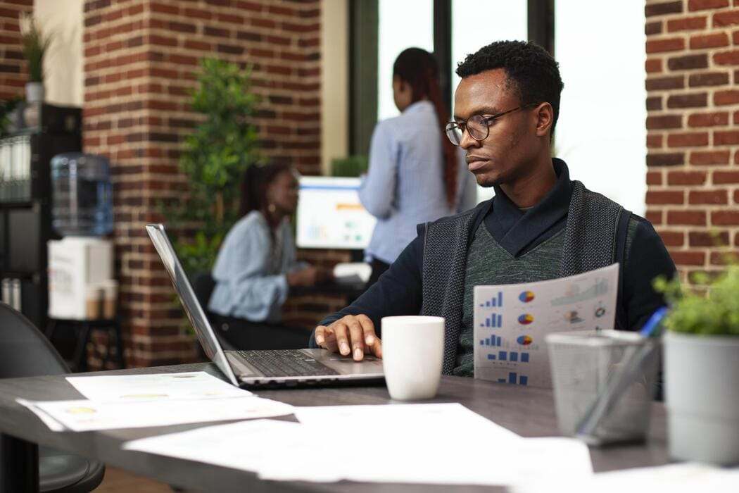Homme à lunettes travaillant sur un ordinateur portable, une page de graphiques imprimée à la main, des collègues derrière lui dans un bureau aux murs de briques