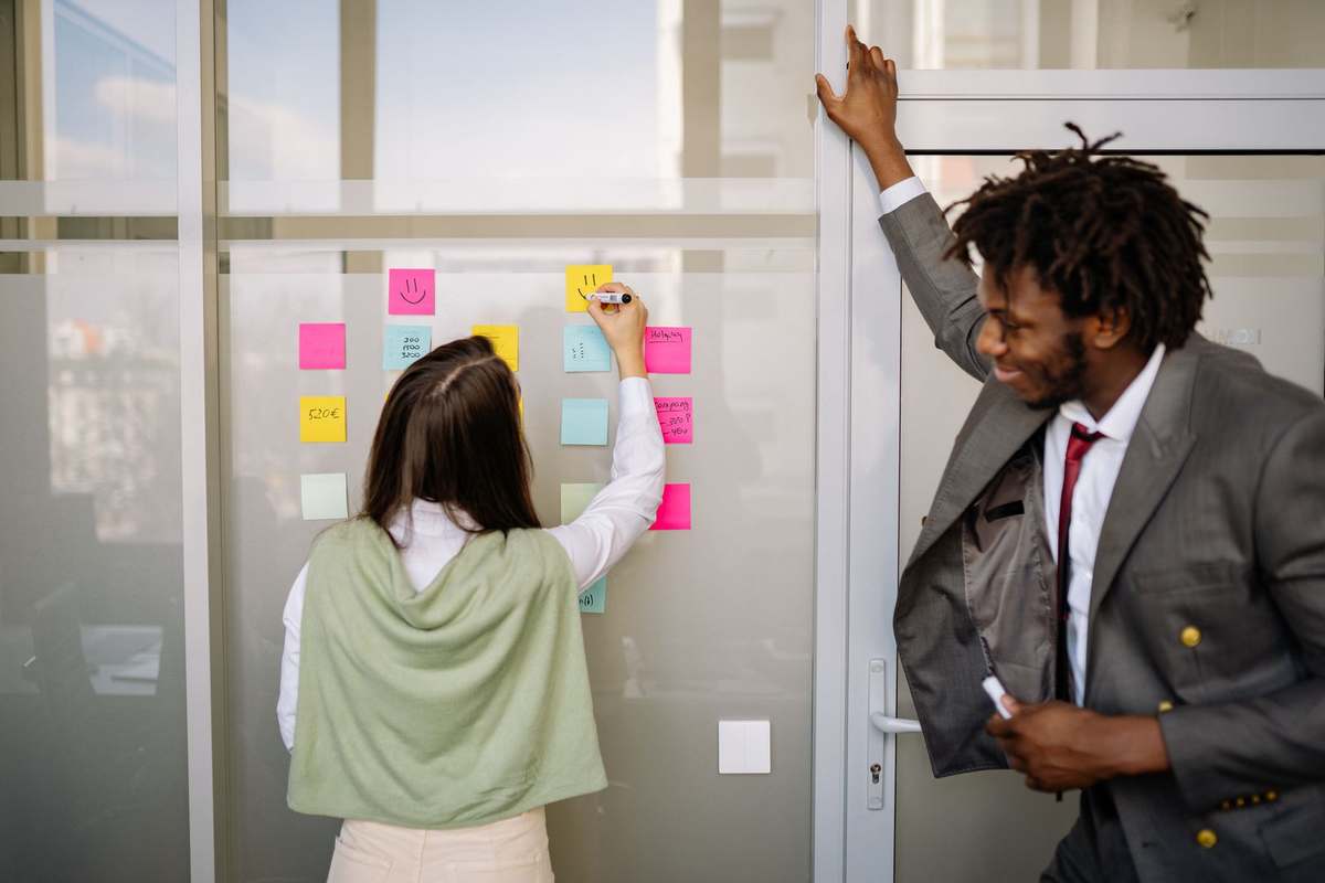 Two colleagues planning a project schedule with sticky notes on a glass office wall
