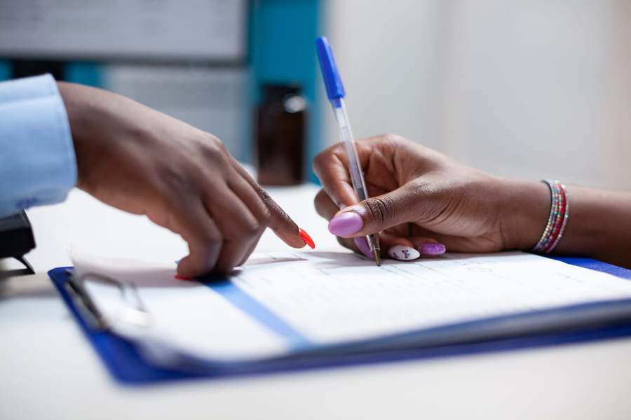 Close-up of two people's hands at a desk: one points to a line on a form on a clipboard while the other fills it in with a pen