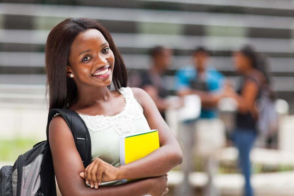 Smiling young woman with a backpack holding a yellow notebook outside a campus building, with other students behind her