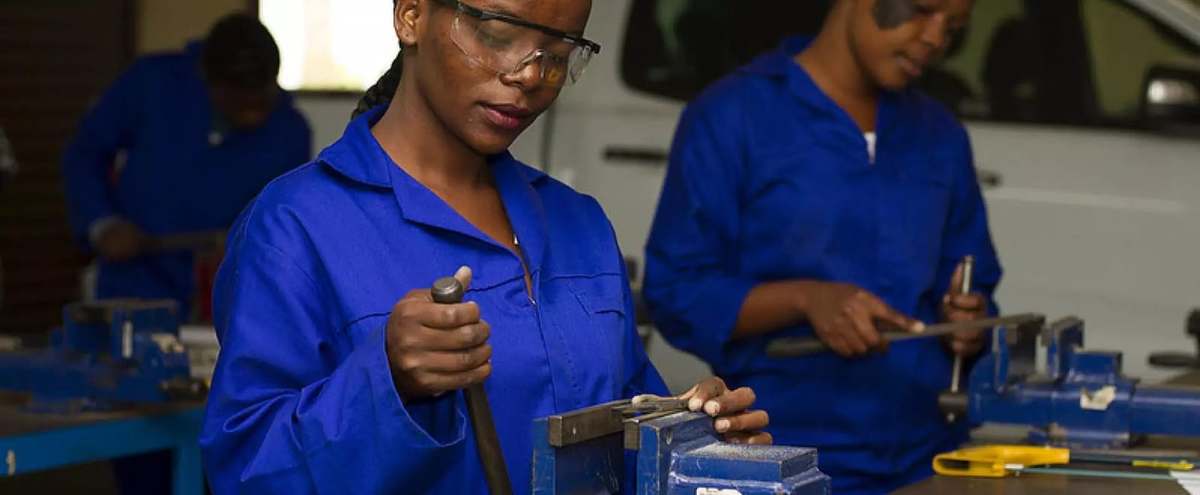Jeune femme en combinaison bleue et lunettes de protection limant une pièce serrée dans un étau d’établi, d’autres apprenants derrière elle
